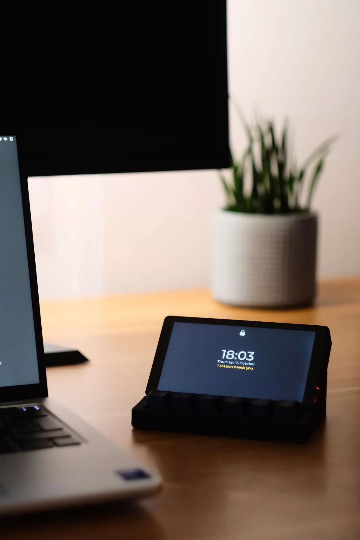 The board in its dock in warm evening light, a plant and a monitor behind it. The lock screen reads 18:03 and 1 session needs you.