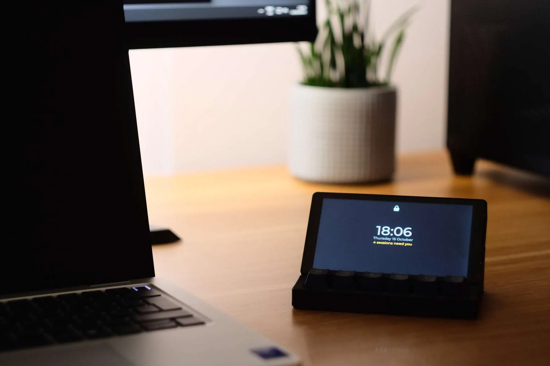 Argus Deck standing in its dock on a wooden desk beside a laptop. Its lock screen reads 18:06, Thursday 15 October, 4 sessions need you.