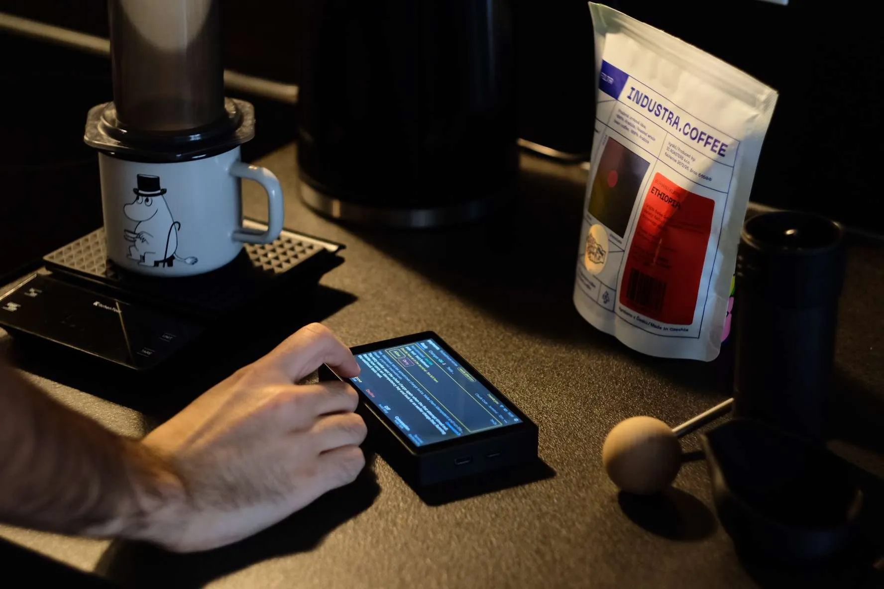 A hand tapping the board on a dark counter, next to a coffee brewer and a bag of beans. The screen shows a permission request.